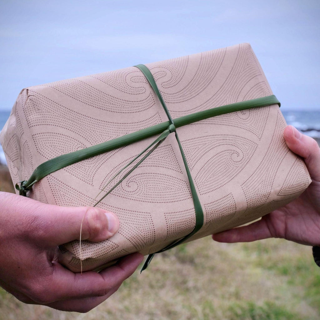 Maori designed paper wrapped package with green flax harakeke ribbon held by hands against a blurred natural background